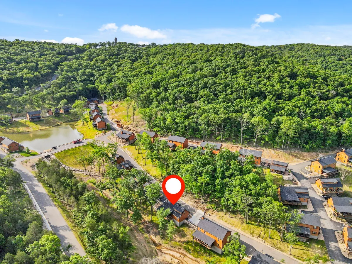 A picturesque view of wooden cabins nestled among lush green trees, with a tranquil lake and winding roads nearby. The landscape invites visitors to explore walking trails, go fishing, or simply relax by the water, embodying the peaceful, nature-filled amenities of the resort.