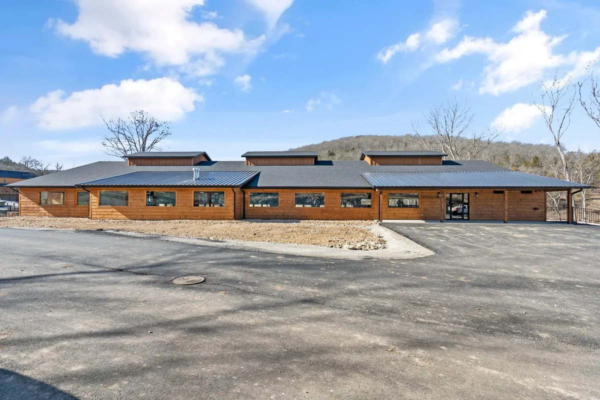 A wide view of Whispering Creek’s main wooden building, featuring large windows that hint at the brand-new interiors, surrounded by lush landscaping and open skies. The setting feels fresh and unfinished, with the sense of a developing, modern community in a natural environment.