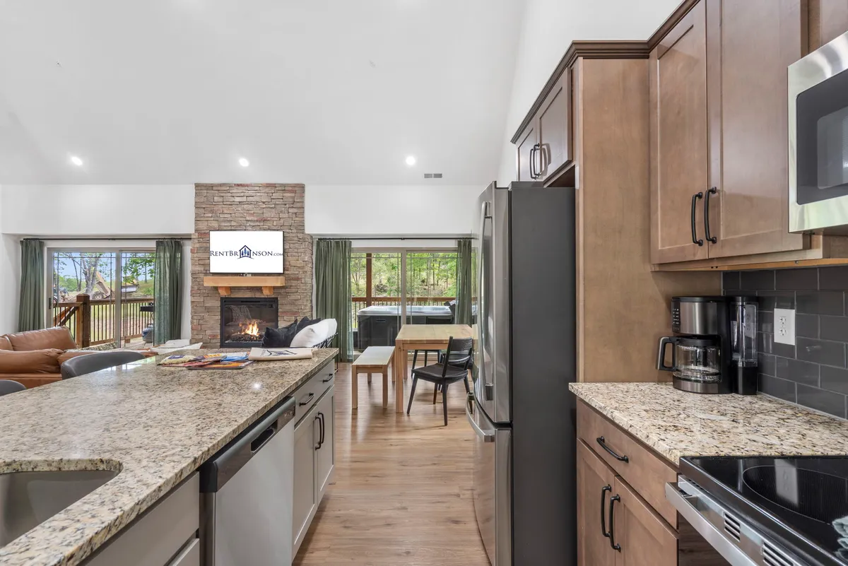 This shot highlights a spacious, inviting living area that seamlessly blends the kitchen, dining, and sitting spaces. A striking stone fireplace anchors the room, with a large dining table and granite countertops emphasizing the modern design and open layout. Expansive windows provide views and natural light.
