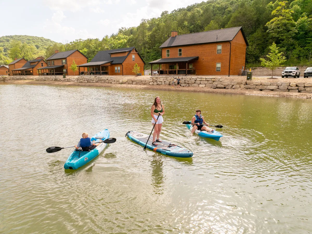A group of kayakers is seen paddling on a serene lake, with cozy cabins and lush trees lining the shore. The setting radiates adventure and togetherness, suggesting opportunities for fishing, boating, and lakeside exploration.