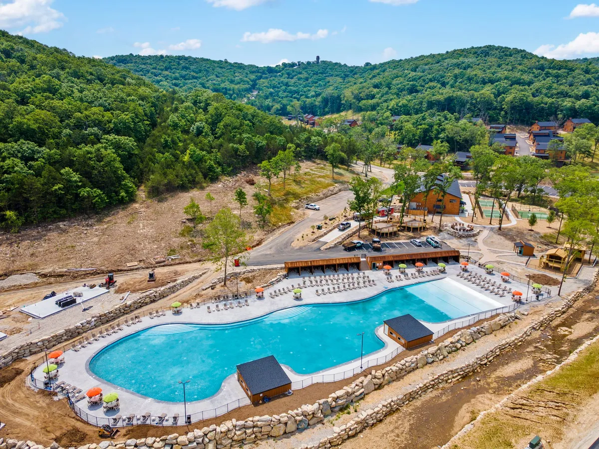 An expansive outdoor swimming pool surrounded by lush greenery, lounge chairs, outdoor structures, and nearby cabins. The scene conveys families swimming, sunbathing, barbecuing, and gathering together for outdoor games, encapsulating a relaxed and joyful resort vibe.