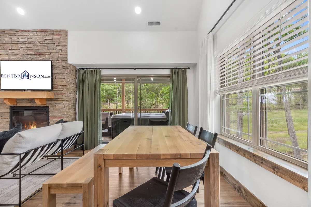 This image captures a stylish open living and dining area with a central dining table, a modern fireplace, large windows that invite natural light, and a television visible in the background. Plush seating and decorative touches set a cozy and welcoming mood, while the arrangement emphasizes togetherness and connection.