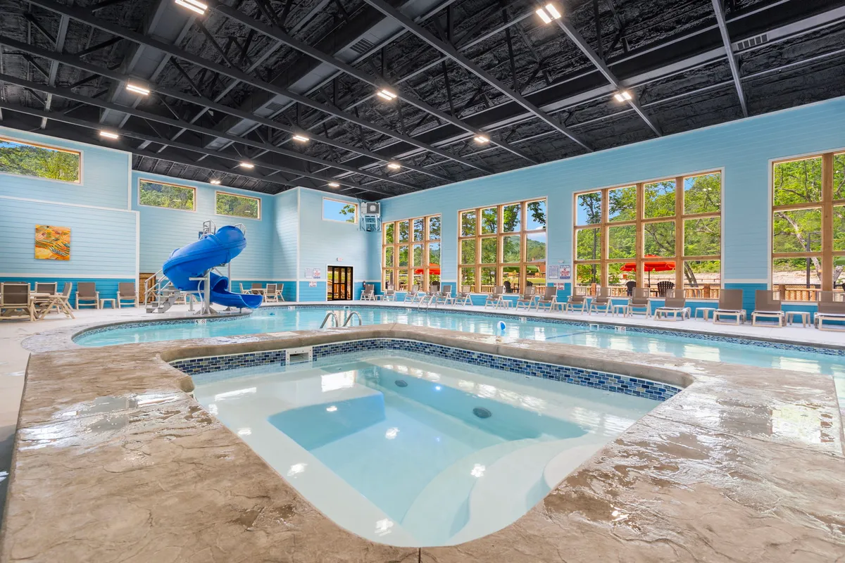 A vibrant indoor pool area featuring a swimming pool, a water slide, a jacuzzi, large windows letting in natural light, and poolside chairs. Children are seen playing on the slide while families relax by the pool, evoking a joyful, family-friendly atmosphere.