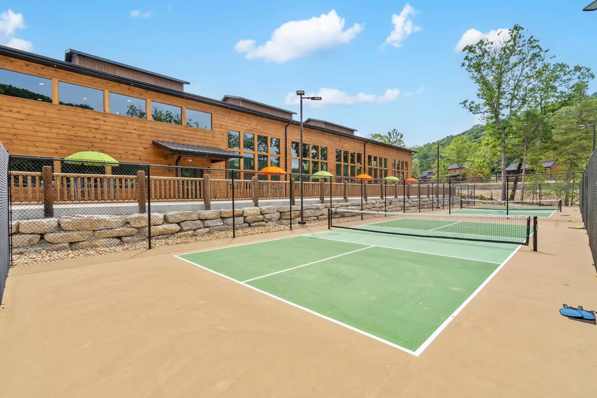 A vibrant shot featuring tennis courts adjacent to a wooden building and outdoor seating areas, with people socializing and relaxing beneath umbrellas. There’s a lively, energetic atmosphere, but the presence of construction or newly finished amenities is subtly implied in the setting.