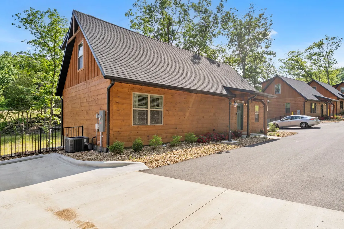 A welcoming exterior scene with a wooden cabin, manicured gardens, a driveway, and mature trees. The image evokes the sense of community growth, outdoor amenities, and natural tranquility, while also suggesting ongoing landscaping and development typical of a new resort.