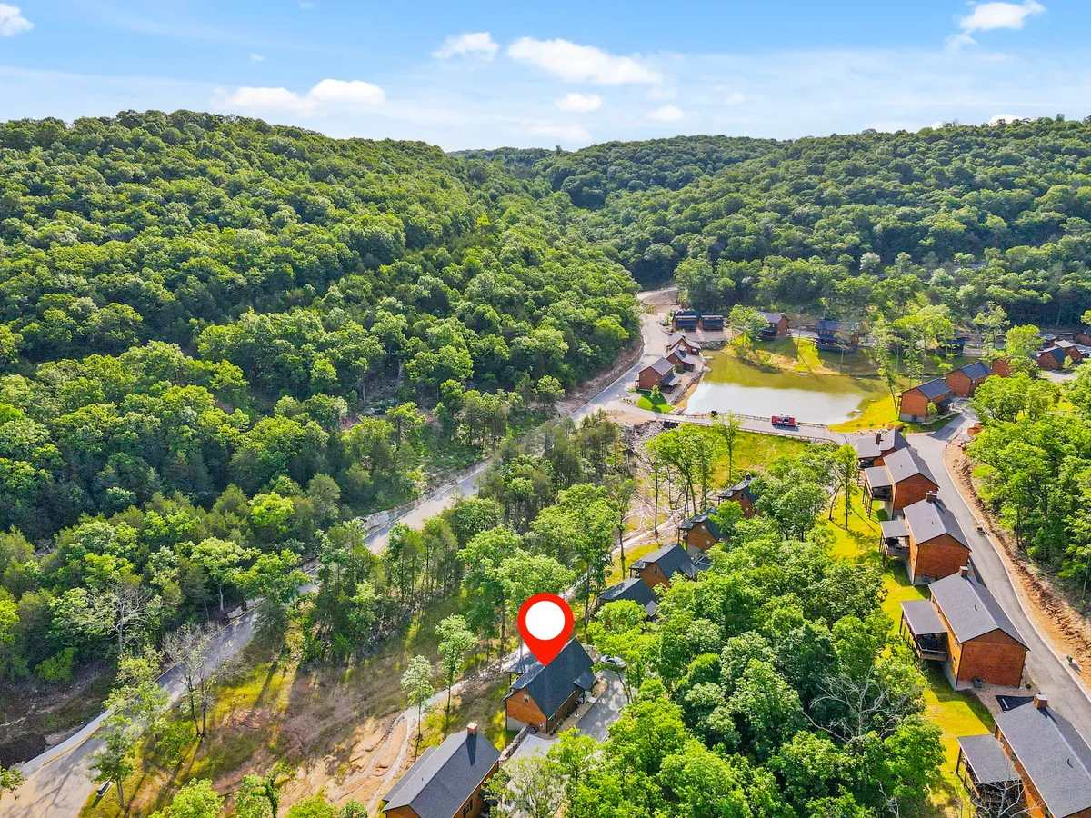 A broad, inviting exterior shot featuring a collection of wooden cabins, lush greenery, a winding road, a reflective pond, and distant scenic hills. The landscape evokes a relaxed, vacation-like atmosphere perfect for families and nature lovers.