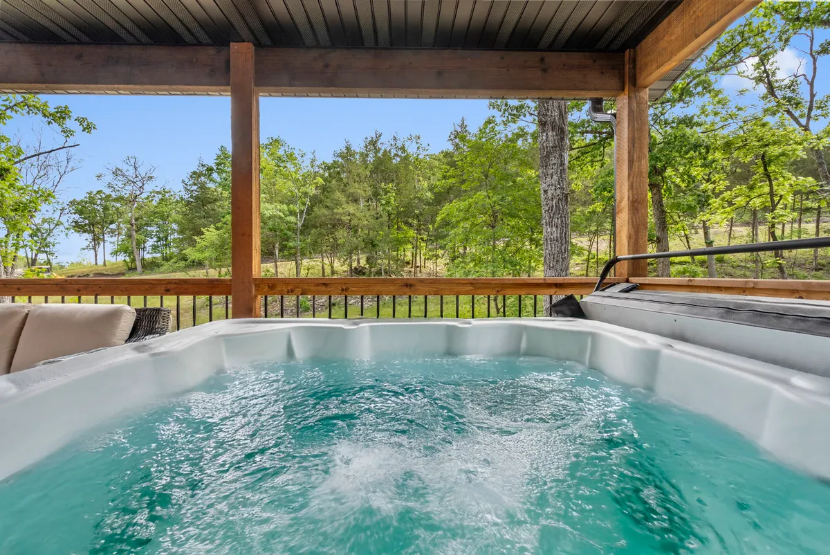 The image showcases the exterior of a charming wooden house nestled among lush greenery, with a private hot tub as the focal point. The setting is tranquil, featuring sunset views over the rolling Ozark hills, inviting outdoor relaxation and socializing amid nature.