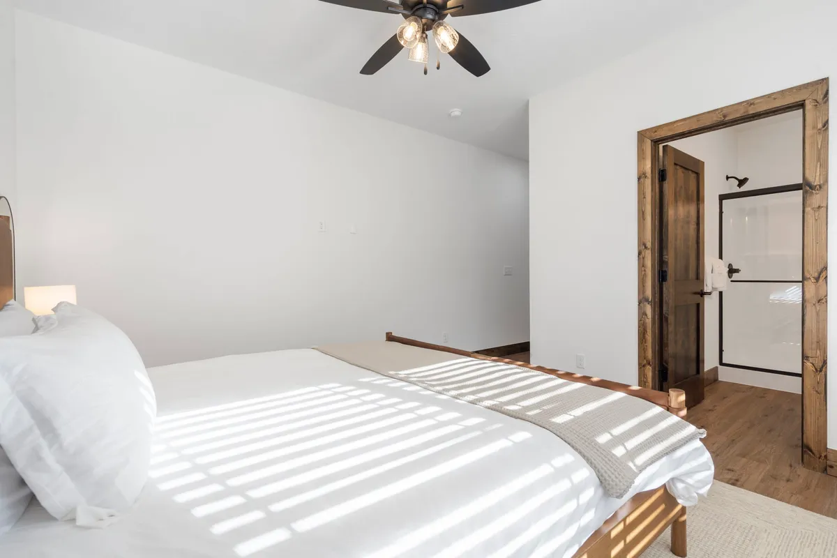 A tranquil king bedroom featuring a bed with crisp white linens, a sturdy wooden frame, a ceiling fan above, and a bathroom door visible in the background. Natural light softly fills the space, highlighting the restful ambiance and modern decor.